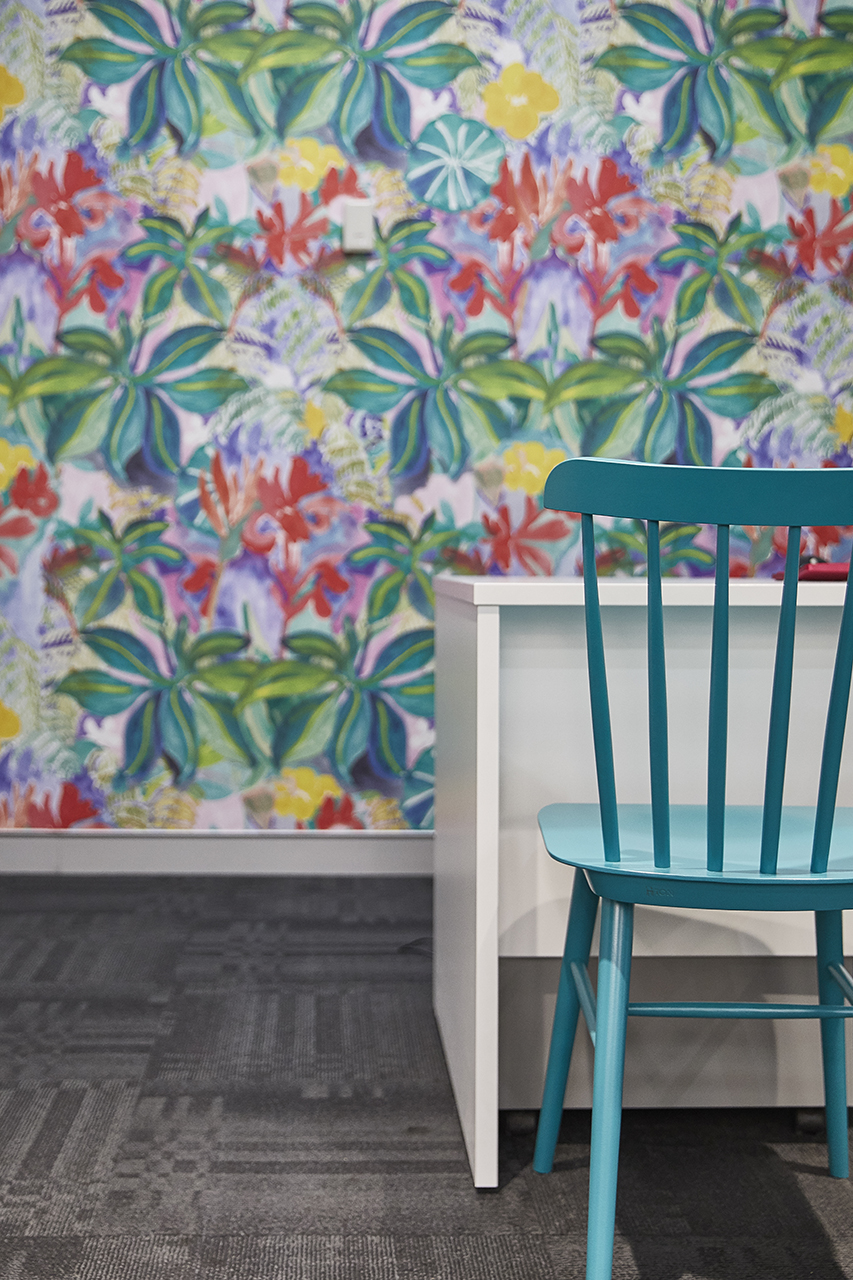 A close-up view of a teal blue wooden chair next to a white desk, set against a wall covered in vibrant, colorful floral and leaf pattern wallpaper and gray patterned carpet.