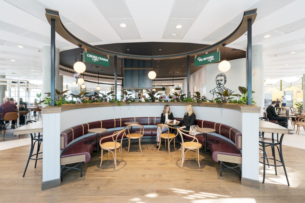 Two women are seated at small circular tables in a semi-circular booth area of a cafe or restaurant. The booth has deep red, upholstered seating and a low wooden wainscoting surrounds the space. The women are engaged in conversation, both holding drinks. The flooring is light wood. In the background, there is a dark ceiling and an upper level or mezzanine with a railing adorned with potted plants. Signs with white text on a green background, reading "CHEERS CHARLIE" and "YES MILORD," are visible above the seating area.