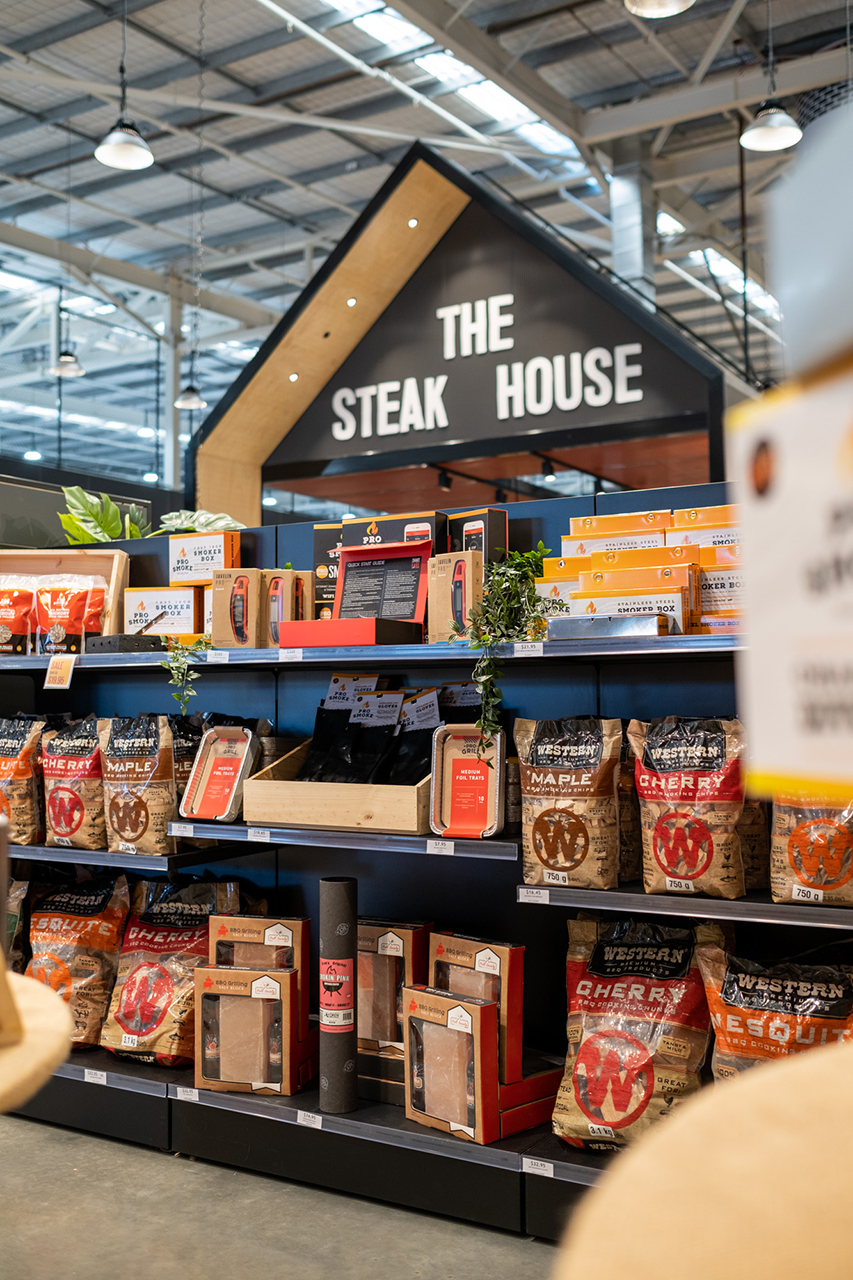 Retail display in a BBQ store with wood chips and sauces under a sign for "THE STEAK HOUSE."