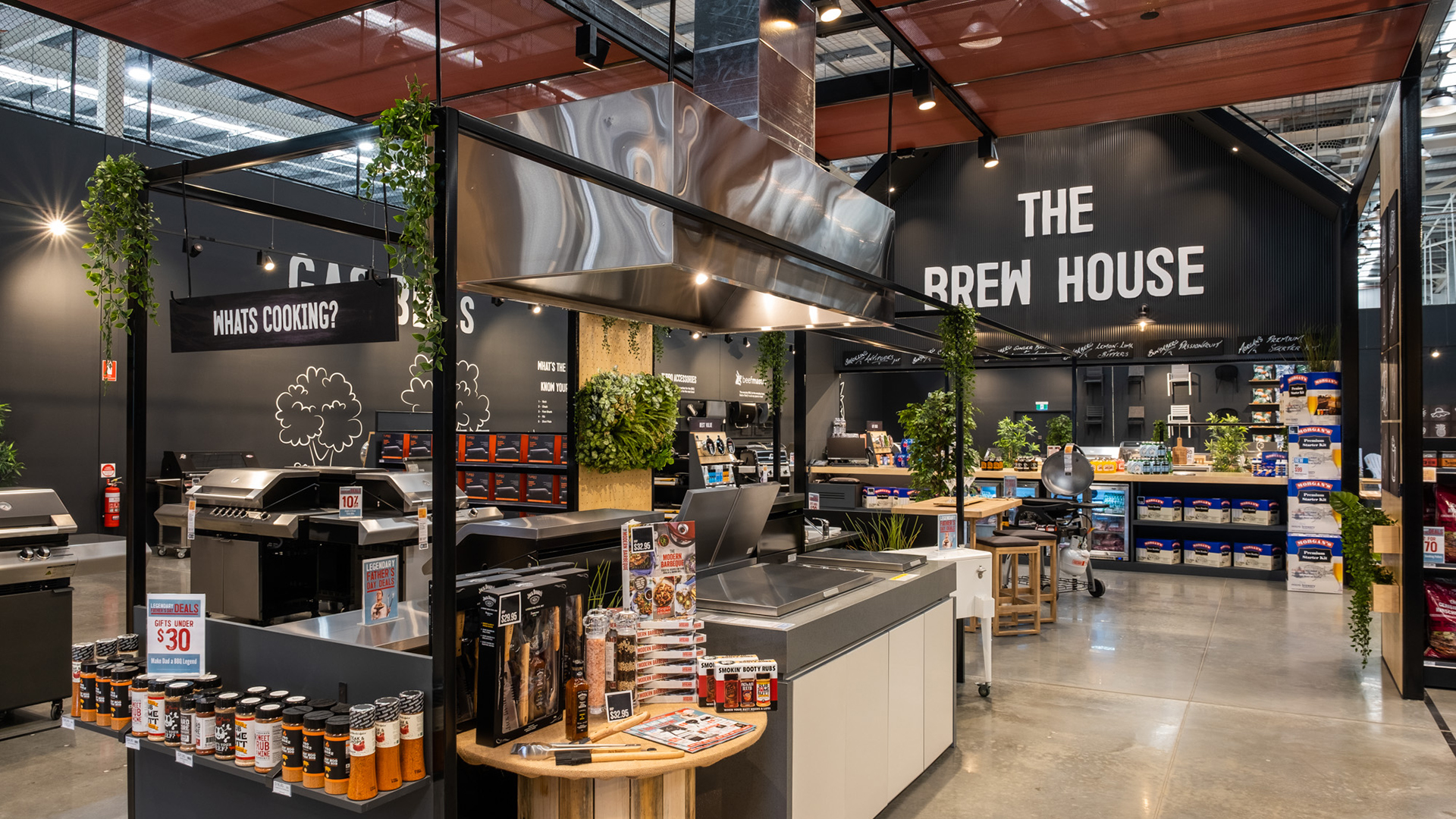 Retail display of an indoor kitchen setup with a large stainless steel range hood, surrounded by grilling products and a sign for "THE BREW HOUSE."