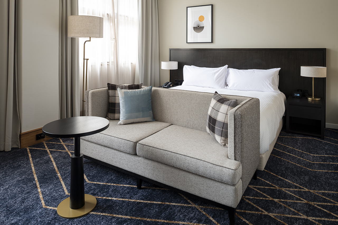 A contemporary hotel room featuring a grey sofa with plaid and blue pillows, a king-size bed with a dark wood headboard, and a round black side table, all set on a geometric navy blue and gold carpet.