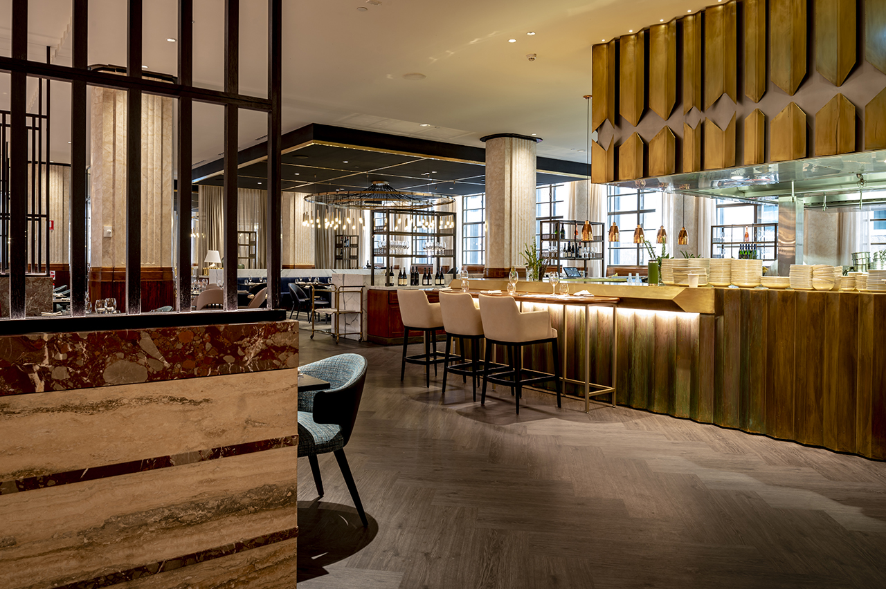 A luxurious hotel restaurant with a warm, dark color palette, featuring a bar with wood and gold accents, beige bar stools, and herringbone wood-look flooring.