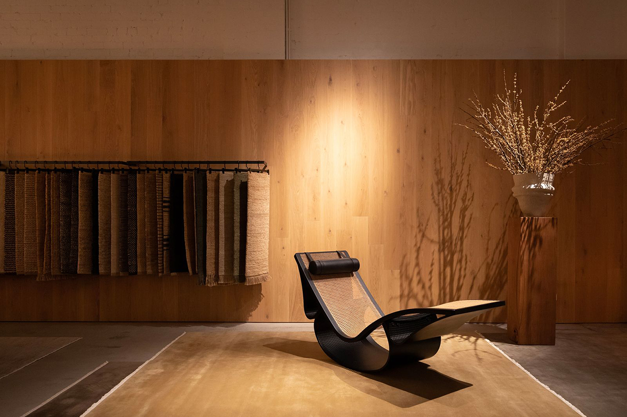 A modern, low-slung chaise lounge with a black frame and woven cane seat is positioned on a large golden-brown rug. In the background, a display of rug samples hangs against a smooth, light wood-paneled wall, complemented by a tall vase of dried branches on a wood pedestal.