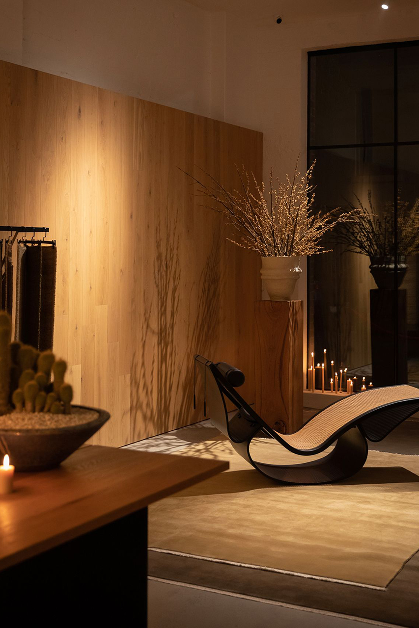 A dimly lit interior space features a modern chaise lounge with a black frame and woven cane surface resting on a rug. A vase of dried branches sits on a wooden pedestal next to a large window, while a bowl of cacti and several lit candles contribute to the warm atmosphere.