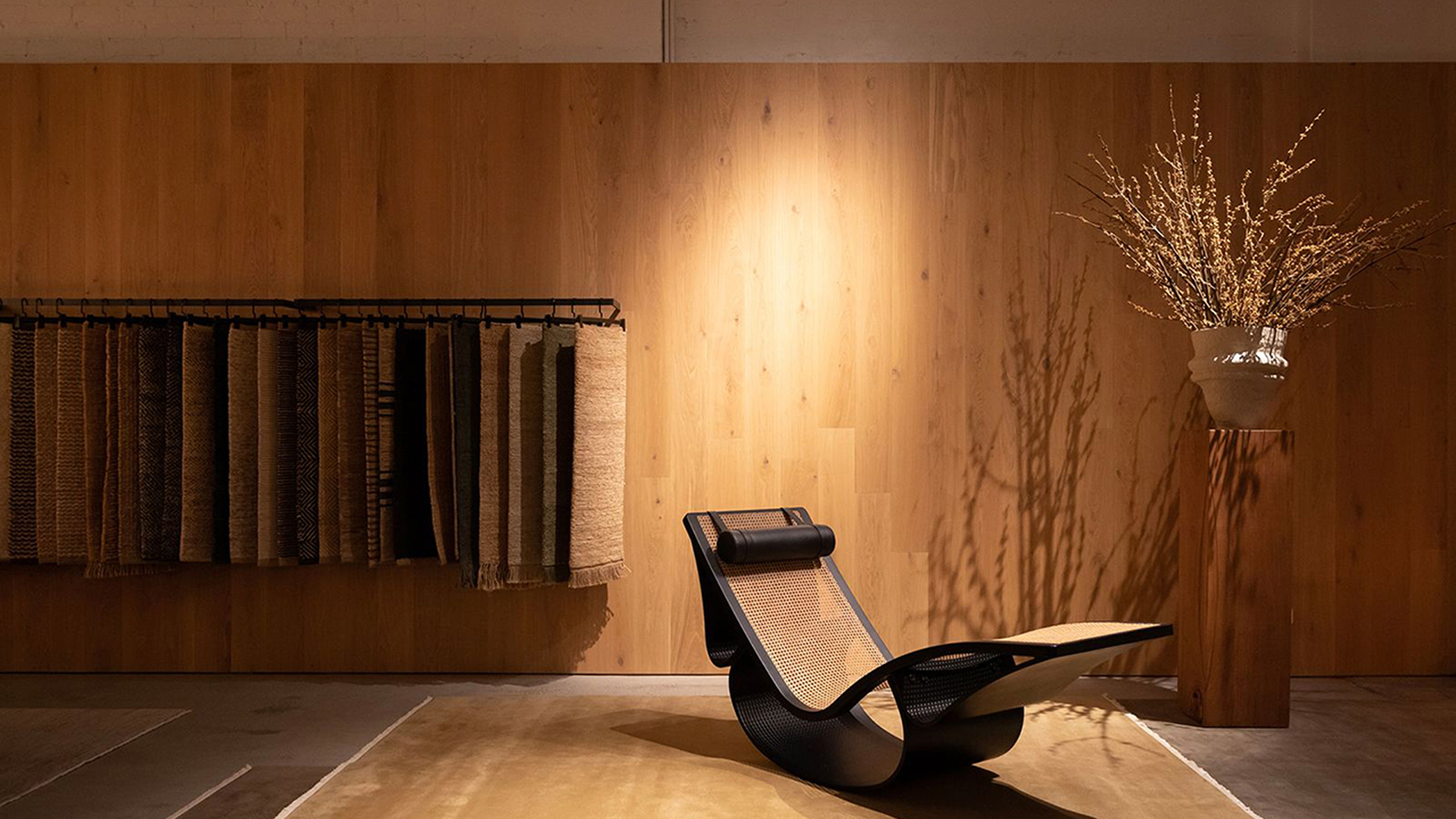 A modern, low-slung chaise lounge with a black frame and woven cane seat is positioned on a large golden-brown rug. In the background, a display of rug samples hangs against a smooth, light wood-paneled wall, complemented by a tall vase of dried branches on a wood pedestal.