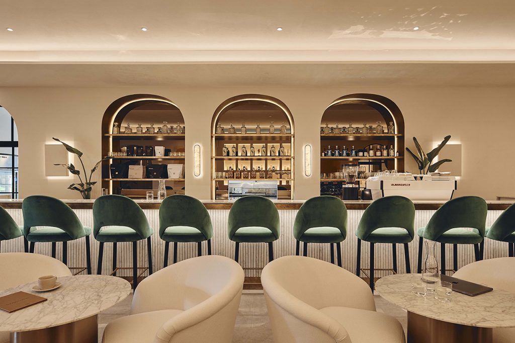 An elegant bar interior featuring green velvet bar stools, arched shelving displaying liquor bottles, and creamy upholstered armchairs in the foreground.