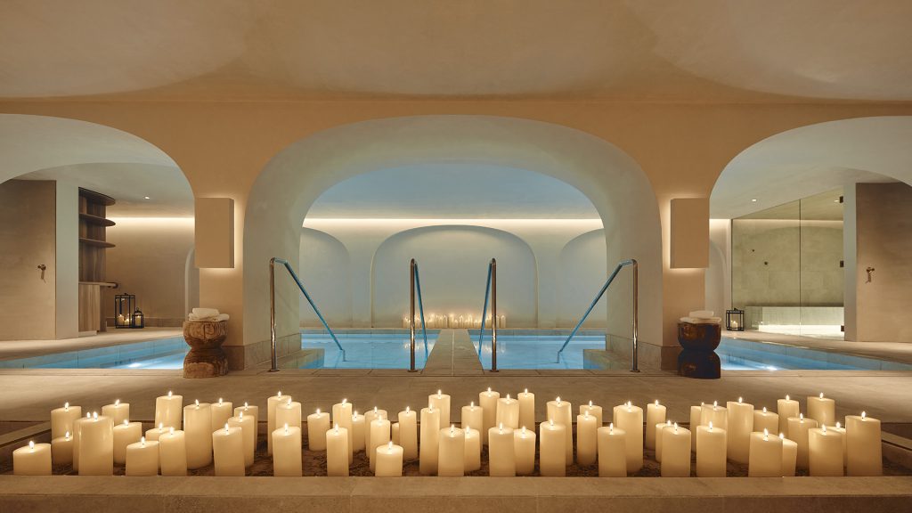 A serene, luxurious spa setting featuring multiple pools under arches, softly lit by strip lighting, and a large display of pillar candles in the foreground.