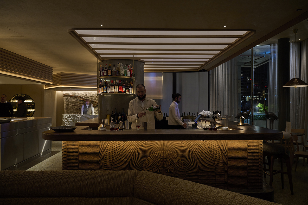 A dimly lit, sophisticated bar with two bartenders working behind a curved wooden bar counter with a textured radial pattern.