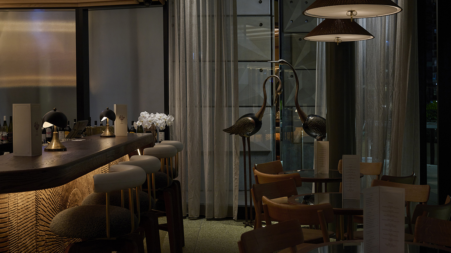 A dimly lit, stylish bar area with a wooden counter, white upholstered stools, and two tall, curved swan sculptures near the windows