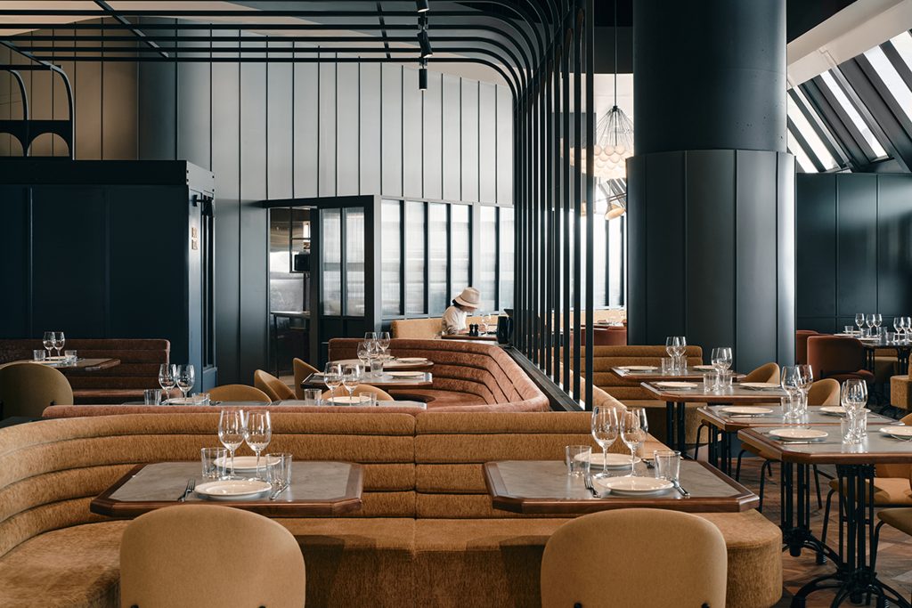 This stylish restaurant dining area features a curved, textured brown banquette and small set tables with a dark metal and fluted wall backdrop.