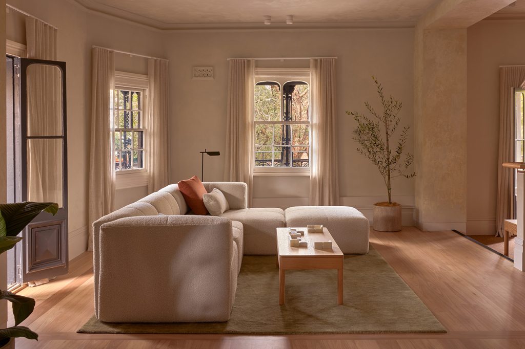 Retail fitout for Eva Surry Hills built by Unita Australia, featuring a white curved fluffy couch on a green rug with a wooden coffee table, framed by old Victorian windows overlooking a green tree and an olive plant in the corner.